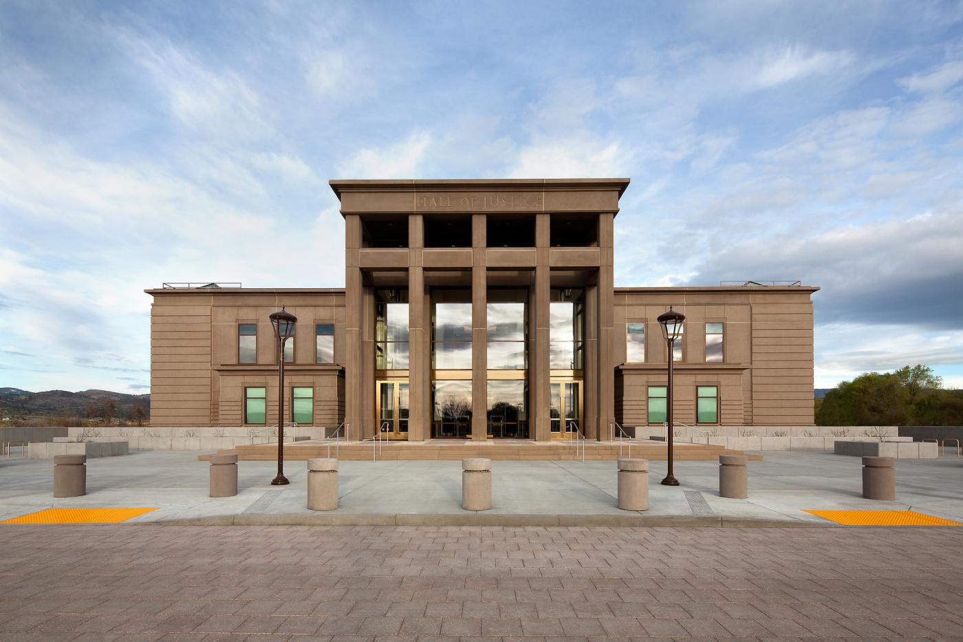 Front exterior of the Lassen County Superior Court Hall of Justice in Susanville, California, a modern tan courthouse with tall central columns and glass entry doors, viewed from the plaza with bollards and lamp posts under a partly cloudy sky.