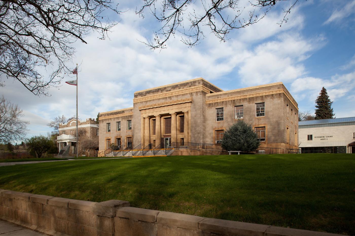 Exterior of Lassen County’s second courthouse in Susanville, California—a two-story stone courthouse with a central columned entrance, viewed from a wide grassy lawn with the U.S. and California flags on a flagpole at left under a partly cloudy sky.
